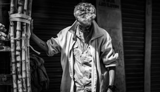 A man with glasses stands next to a bundle of sugarcanes in a contemplative pose, wearing a jacket and looking down. The setting appears to be an urban street with closed shutters in the background. The image is in black and white, highlighting shadows and textures.
