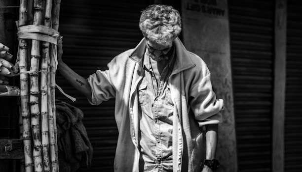 A man with glasses stands next to a bundle of sugarcanes in a contemplative pose, wearing a jacket and looking down. The setting appears to be an urban street with closed shutters in the background. The image is in black and white, highlighting shadows and textures.