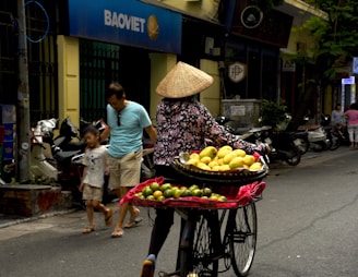 A street vendor wearing a conical hat rides a bicycle equipped with baskets filled with yellow and green fruits. A man and child walk nearby, passing a row of parked motorcycles. The setting is an urban street with buildings, including a shop or bank with a sign labeled 'BAOVIET'. Trees and pedestrians are visible in the background.