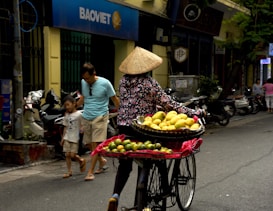 A street vendor wearing a conical hat rides a bicycle equipped with baskets filled with yellow and green fruits. A man and child walk nearby, passing a row of parked motorcycles. The setting is an urban street with buildings, including a shop or bank with a sign labeled 'BAOVIET'. Trees and pedestrians are visible in the background.