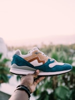 A hand holds a stylish sneaker against a soft, blurred background of palm trees. The shoe features a blue and tan suede design with white mesh accents.