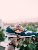 A hand holds a stylish sneaker against a soft, blurred background of palm trees. The shoe features a blue and tan suede design with white mesh accents.