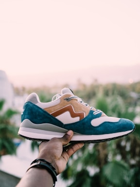 A hand holds a stylish sneaker against a soft, blurred background of palm trees. The shoe features a blue and tan suede design with white mesh accents.