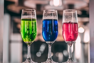 Three tall glass flutes filled with colorful liquids are arranged in a row. The liquid in the left glass is bright green, the middle glass contains a vivid blue liquid, and the right glass holds a deep pink liquid. The background is blurred, suggesting an indoor setting, possibly a bar or restaurant, with soft lighting that creates a cozy atmosphere.