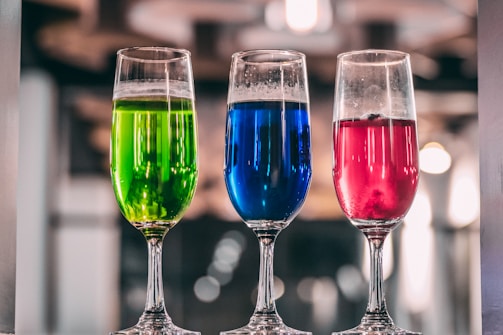 Three tall glass flutes filled with colorful liquids are arranged in a row. The liquid in the left glass is bright green, the middle glass contains a vivid blue liquid, and the right glass holds a deep pink liquid. The background is blurred, suggesting an indoor setting, possibly a bar or restaurant, with soft lighting that creates a cozy atmosphere.