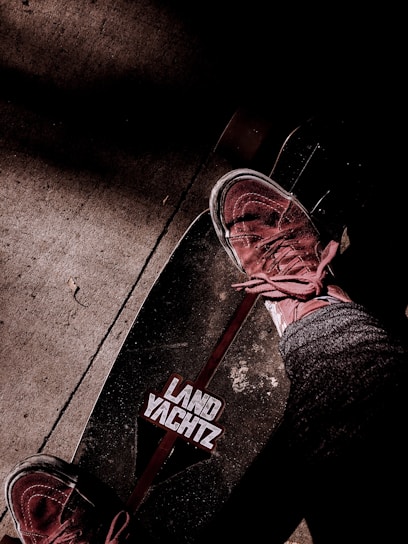 A skateboard with the brand 'Land Yachtz' visible, accompanied by a person wearing sneakers and jeans standing on it. The background features a textured concrete surface with shadows adding depth to the scene.