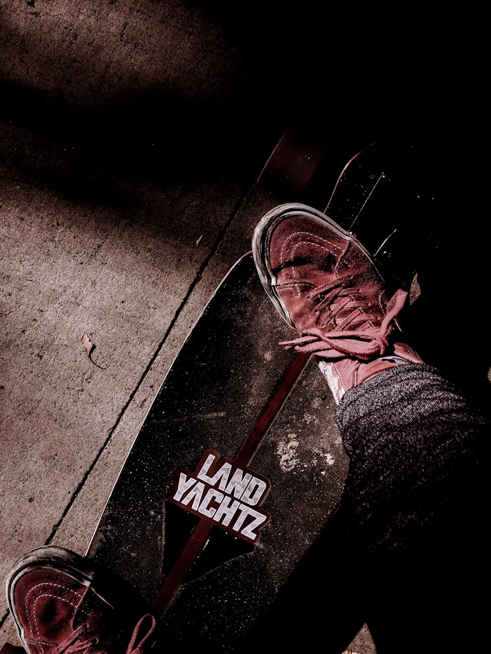 A skateboard with the brand 'Land Yachtz' visible, accompanied by a person wearing sneakers and jeans standing on it. The background features a textured concrete surface with shadows adding depth to the scene.