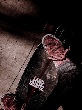 A skateboard with the brand 'Land Yachtz' visible, accompanied by a person wearing sneakers and jeans standing on it. The background features a textured concrete surface with shadows adding depth to the scene.