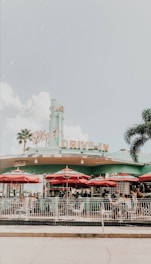A retro-style drive-in diner with teal and pink colors, featuring a classic neon sign reading 'Drive-In'. Several red umbrellas shade outdoor tables where people are seated, surrounded by palm trees and a clear blue sky.