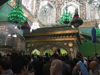 A crowded indoor scene with ornate, gold, and intricate architectural details, including decorative elements and Arabic calligraphy. Large, green chandeliers hang from the ceiling, casting a green glow. The focal point is a beautifully decorated shrine with floral arrangements on top. A large group of people gathers around, some reaching out, signaling a religious or cultural gathering.