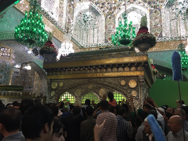 A crowded indoor scene with ornate, gold, and intricate architectural details, including decorative elements and Arabic calligraphy. Large, green chandeliers hang from the ceiling, casting a green glow. The focal point is a beautifully decorated shrine with floral arrangements on top. A large group of people gathers around, some reaching out, signaling a religious or cultural gathering.
