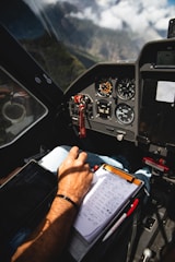 The image captures the cockpit interior of an aircraft with a focus on the control panel and instrumentation. A hand is seen writing on a checklist or notes, indicating active monitoring or calculations. The view outside the cockpit window shows a mountainous landscape under a partly cloudy sky.