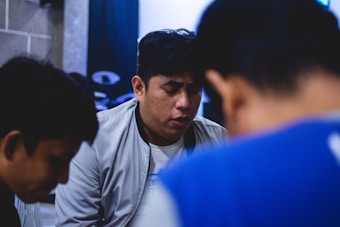 A group of three people is sitting closely together, with one person in the center wearing a light-colored jacket, appearing deep in thought or prayer. The background is slightly blurred and includes a drum set.