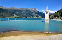 A picturesque scene featuring a submerged bell tower rising from a tranquil turquoise lake, surrounded by lush green hills and rugged mountains under a clear blue sky. A pathway with several people is visible on the far side of the lake.