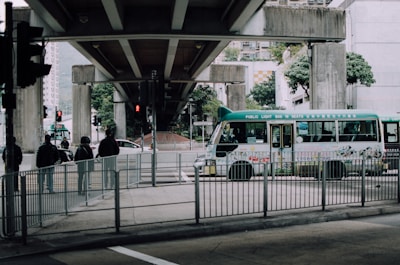 A green public light bus is passing under a concrete overpass, surrounded by urban structures and trees. Several pedestrians wearing jackets are standing near metal railings while traffic lights are visible in the background.