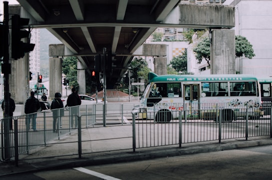 A green public light bus is passing under a concrete overpass, surrounded by urban structures and trees. Several pedestrians wearing jackets are standing near metal railings while traffic lights are visible in the background.
