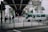 A green public light bus is passing under a concrete overpass, surrounded by urban structures and trees. Several pedestrians wearing jackets are standing near metal railings while traffic lights are visible in the background.