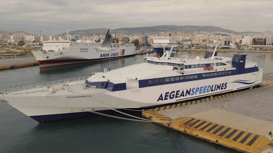 Two large ferries are docked at a port, with a cityscape and hills in the background. One ship has 'Anek Lines' written on it, and the other features 'Aegean Speedlines'. The water is calm, and the sky is overcast.