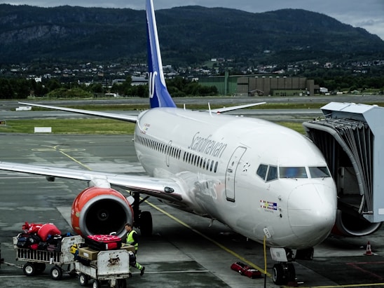 A commercial airplane is parked at an airport gate, with a jet bridge connected to the aircraft. It is a Scandinavian Airlines plane, and there is luggage being loaded onto it by ground crew. The surroundings include an airfield with mountains and green landscape in the background, along with cloudy skies.