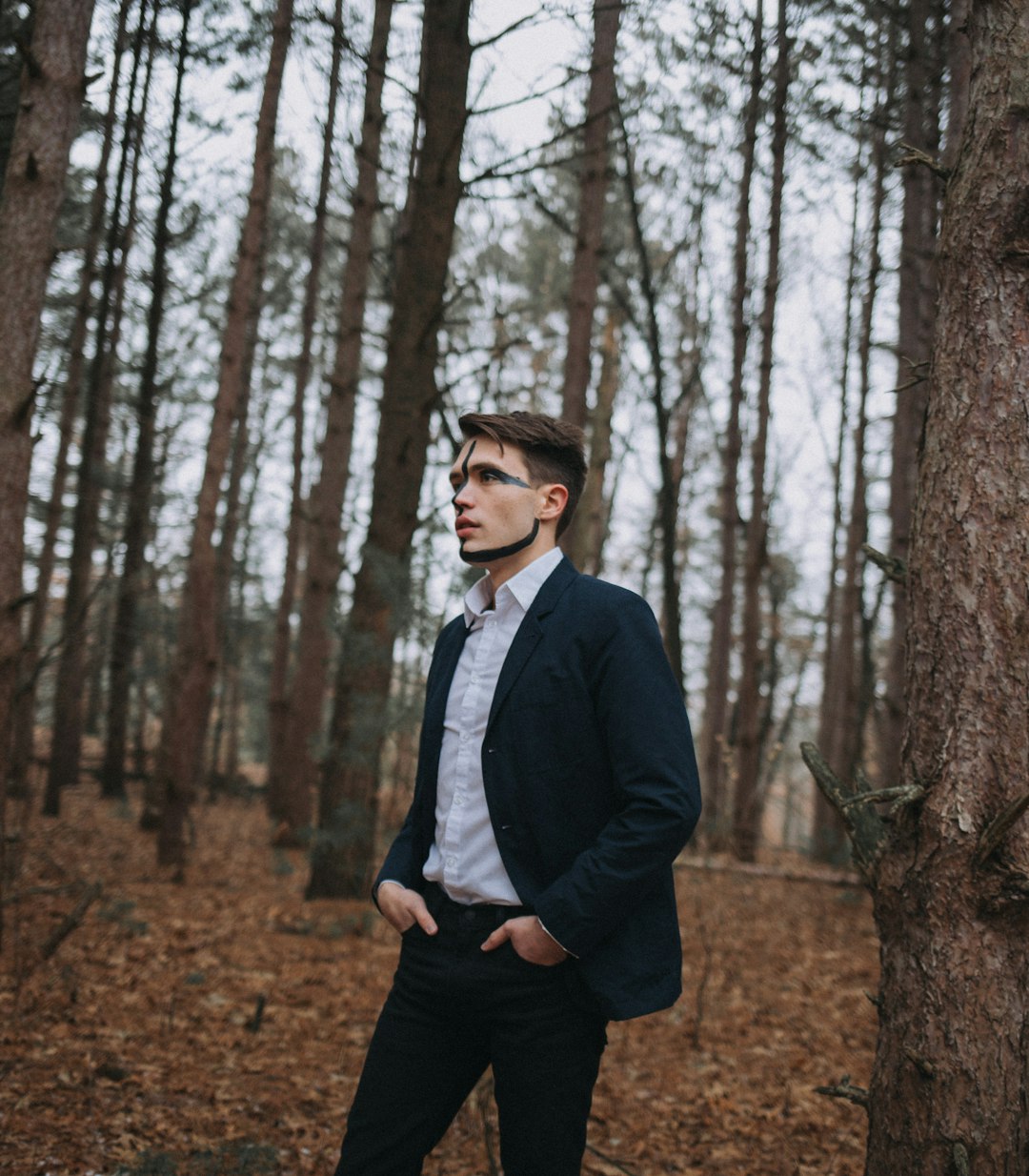 man in blue blazer and black pants stands beside tall trees