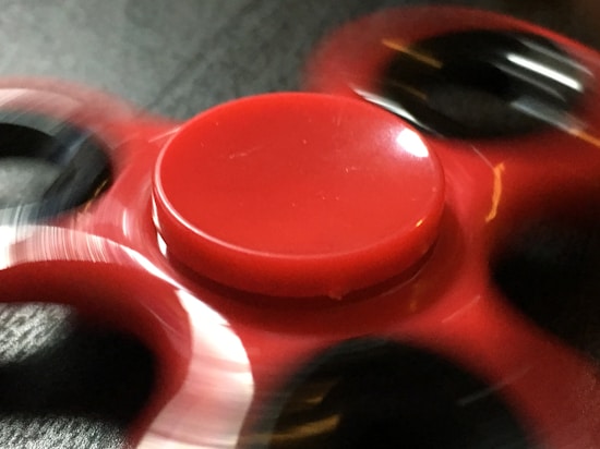 A red fidget spinner in motion, causing blur effects around its central hub and the three circular lobes. The background is dark, highlighting the bright red color and movement.