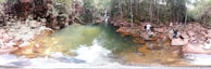 A natural pool surrounded by rocky terrain and lush greenery, with a small waterfall in the background. Several people are sitting and standing on the rocks, some dipping their feet into the water. The setting is serene and appears to be a popular spot for relaxation and enjoying nature.