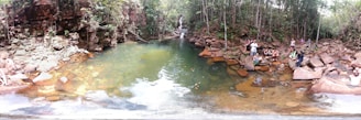 A natural pool surrounded by rocky terrain and lush greenery, with a small waterfall in the background. Several people are sitting and standing on the rocks, some dipping their feet into the water. The setting is serene and appears to be a popular spot for relaxation and enjoying nature.