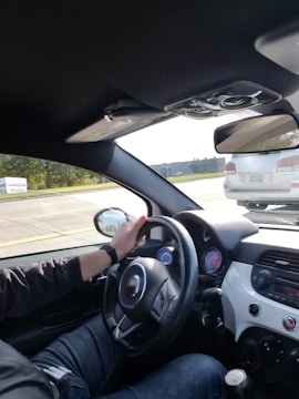The view is from inside a car, showing the driver holding the steering wheel with one hand. The dashboard and controls are visible, and there is a car outside through the window.