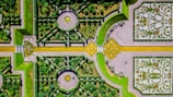 An intricately designed formal garden viewed from above, featuring symmetrical patterns with hedges, pathways, and decorative elements. The garden includes circular and rectangular sections, with yellow and green hues dominating the design. A few people can be seen walking through the garden, and decorative floral patterns enhance the visual appeal.