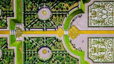 An intricately designed formal garden viewed from above, featuring symmetrical patterns with hedges, pathways, and decorative elements. The garden includes circular and rectangular sections, with yellow and green hues dominating the design. A few people can be seen walking through the garden, and decorative floral patterns enhance the visual appeal.