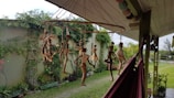 A collection of handmade, rustic doll-like figures made from natural materials hang from a circular mobile outside, with a garden and greenery in the background. The mobile is suspended from the roof of a porch, next to a red hammock. There is a wall covered in climbing plants and various potted plants in the garden.