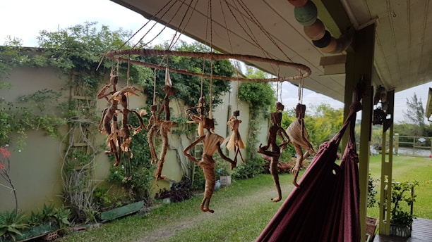A collection of handmade, rustic doll-like figures made from natural materials hang from a circular mobile outside, with a garden and greenery in the background. The mobile is suspended from the roof of a porch, next to a red hammock. There is a wall covered in climbing plants and various potted plants in the garden.