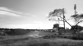 A rural landscape with a dirt road, tall metal structures resembling cell towers, and a building surrounded by fields and trees. Piles of gravel or earth are seen in the distance.