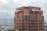 A tall building with a retro-futuristic design has the words 'Hotel Kintetsu' displayed in stylized fonts on its top. The architecture features a combination of red and greenish-brown hues with geometric patterns and circular windows. The view includes industrial areas and a body of water in the background under an overcast sky.