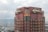 A tall building with a retro-futuristic design has the words 'Hotel Kintetsu' displayed in stylized fonts on its top. The architecture features a combination of red and greenish-brown hues with geometric patterns and circular windows. The view includes industrial areas and a body of water in the background under an overcast sky.