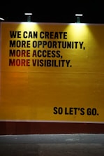 A large, bold statement on a yellow background conveys a motivational message. The text includes the phrases 'We can create more opportunity, more access, more visibility' in capital letters. The words 'So let's go.' are placed separately in the lower right corner, also in capital letters. The text is illuminated by lights from above, casting gentle shadows around the letters, enhancing the commanding and inspirational tone of the message.