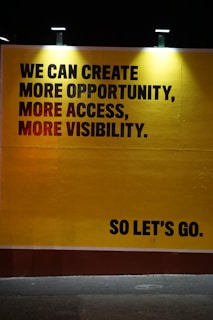A large, bold statement on a yellow background conveys a motivational message. The text includes the phrases 'We can create more opportunity, more access, more visibility' in capital letters. The words 'So let's go.' are placed separately in the lower right corner, also in capital letters. The text is illuminated by lights from above, casting gentle shadows around the letters, enhancing the commanding and inspirational tone of the message.