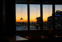 A cityscape is visible through large windows, with a vibrant sunset casting warm orange and yellow tones over the scene. The silhouette of buildings and a bridge is prominent, and the interior has pendant lights hanging down. Dim lighting creates a cozy indoor atmosphere with a person sitting at a table.