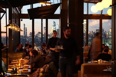 A dimly lit restaurant interior with large windows showcasing a city skyline at dusk. Multiple patrons are seated at tables, enjoying their meals, while a waiter moves through the space. The soft glow of pendant lights and candles on the tables adds warmth to the scene.