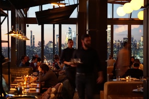 A dimly lit restaurant interior with large windows showcasing a city skyline at dusk. Multiple patrons are seated at tables, enjoying their meals, while a waiter moves through the space. The soft glow of pendant lights and candles on the tables adds warmth to the scene.