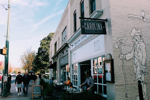 A bustling street scene featuring a coffee shop with outdoor seating on the sidewalk. Several people are having a conversation or walking by. The shop sign reads 'Carolina Coffee Shop' and there's a mural on the wall depicting a historical figure holding a cane and a rose. A chalkboard on the sidewalk advertises mimosas. The setting suggests a lively, community-oriented atmosphere.