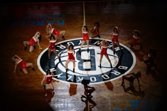 A group of cheerleaders perform in coordinated red outfits around a central basketball team logo on a polished wooden court. The dancers are positioned in dynamic, synchronized poses, suggesting energetic choreography and a lively performance setting.