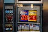An advertisement display in a store window features lottery jackpot amounts for Mega Millions and Powerball, both showing jackpots of 208 million and 200 million respectively. Below the display, several lottery tickets and promotional posters are visible. To the left of the display, there is a refrigerator stocked with various drinks including different flavors of sports drinks and bottled water.