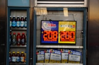 An advertisement display in a store window features lottery jackpot amounts for Mega Millions and Powerball, both showing jackpots of 208 million and 200 million respectively. Below the display, several lottery tickets and promotional posters are visible. To the left of the display, there is a refrigerator stocked with various drinks including different flavors of sports drinks and bottled water.