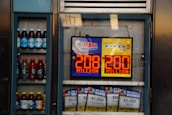 An advertisement display in a store window features lottery jackpot amounts for Mega Millions and Powerball, both showing jackpots of 208 million and 200 million respectively. Below the display, several lottery tickets and promotional posters are visible. To the left of the display, there is a refrigerator stocked with various drinks including different flavors of sports drinks and bottled water.