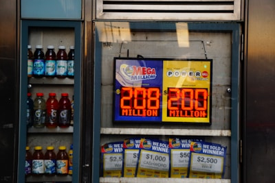 An advertisement display in a store window features lottery jackpot amounts for Mega Millions and Powerball, both showing jackpots of 208 million and 200 million respectively. Below the display, several lottery tickets and promotional posters are visible. To the left of the display, there is a refrigerator stocked with various drinks including different flavors of sports drinks and bottled water.