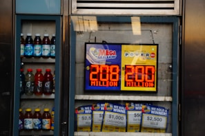 An advertisement display in a store window features lottery jackpot amounts for Mega Millions and Powerball, both showing jackpots of 208 million and 200 million respectively. Below the display, several lottery tickets and promotional posters are visible. To the left of the display, there is a refrigerator stocked with various drinks including different flavors of sports drinks and bottled water.