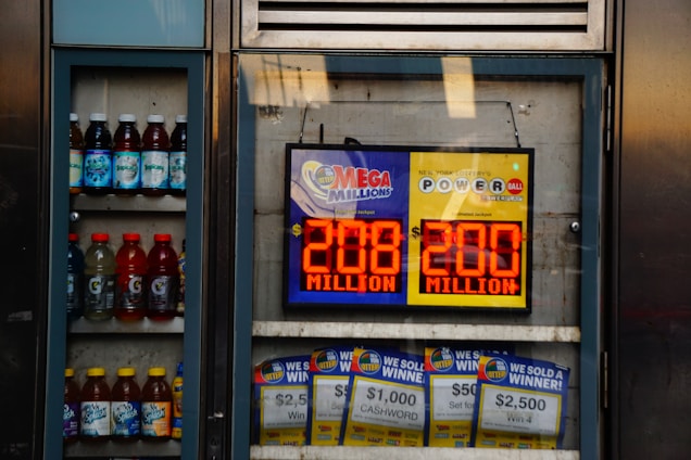 An advertisement display in a store window features lottery jackpot amounts for Mega Millions and Powerball, both showing jackpots of 208 million and 200 million respectively. Below the display, several lottery tickets and promotional posters are visible. To the left of the display, there is a refrigerator stocked with various drinks including different flavors of sports drinks and bottled water.