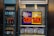 An advertisement display in a store window features lottery jackpot amounts for Mega Millions and Powerball, both showing jackpots of 208 million and 200 million respectively. Below the display, several lottery tickets and promotional posters are visible. To the left of the display, there is a refrigerator stocked with various drinks including different flavors of sports drinks and bottled water.