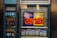 An advertisement display in a store window features lottery jackpot amounts for Mega Millions and Powerball, both showing jackpots of 208 million and 200 million respectively. Below the display, several lottery tickets and promotional posters are visible. To the left of the display, there is a refrigerator stocked with various drinks including different flavors of sports drinks and bottled water.